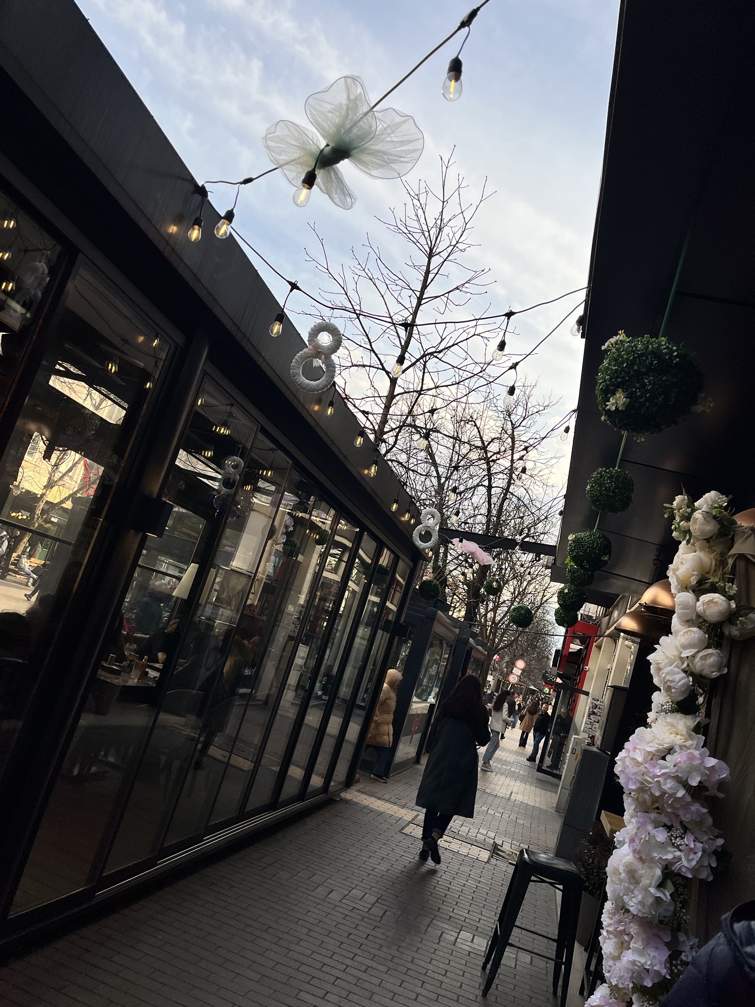 Narrow pedestrian street in Sofia lined with cafés, with string lights overhead and people walking between glass-fronted buildings