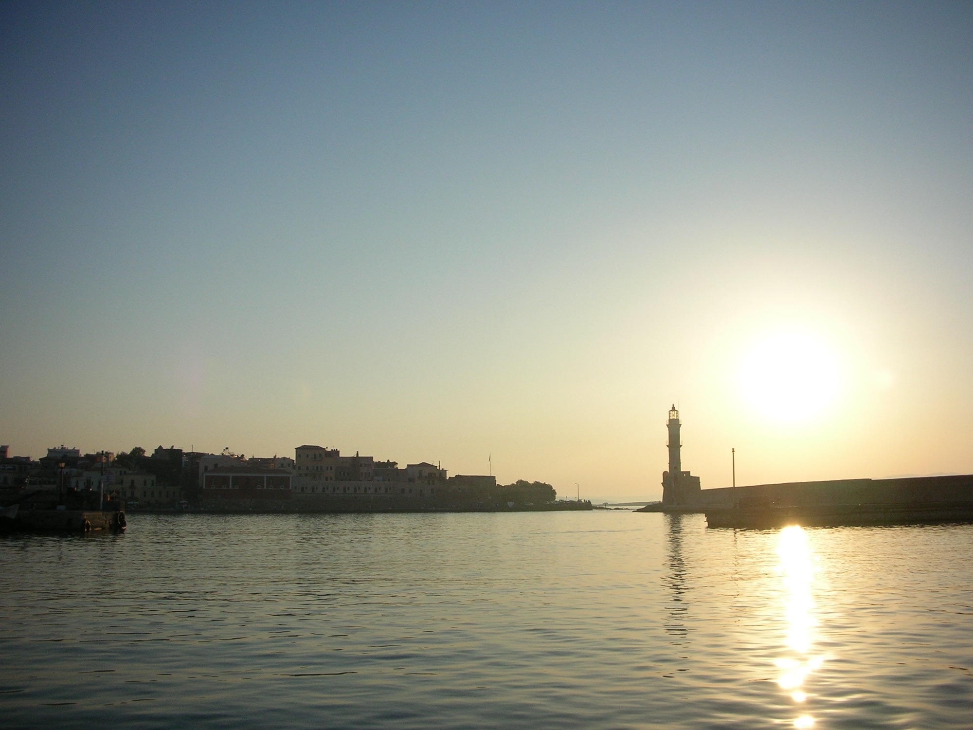 Chania lighthouse silhouetted against a golden sunset, its reflection shimmering on the calm harbour water